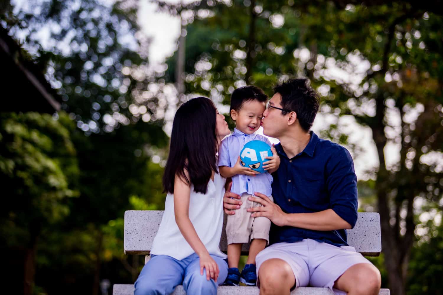 family photoshoot singapore a mother and father sitting on a bench and kissing on the cheeks of their adorable son
