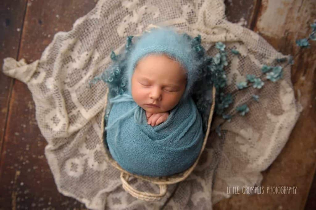 baby photoshoot Londonnewborn is placed in a basket with bunch of blue flowers and covered with blue blanket